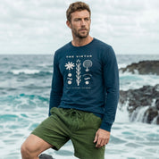 Man wearing a blue shirt and green shorts standing on a rocky beach with ocean waves in the background.