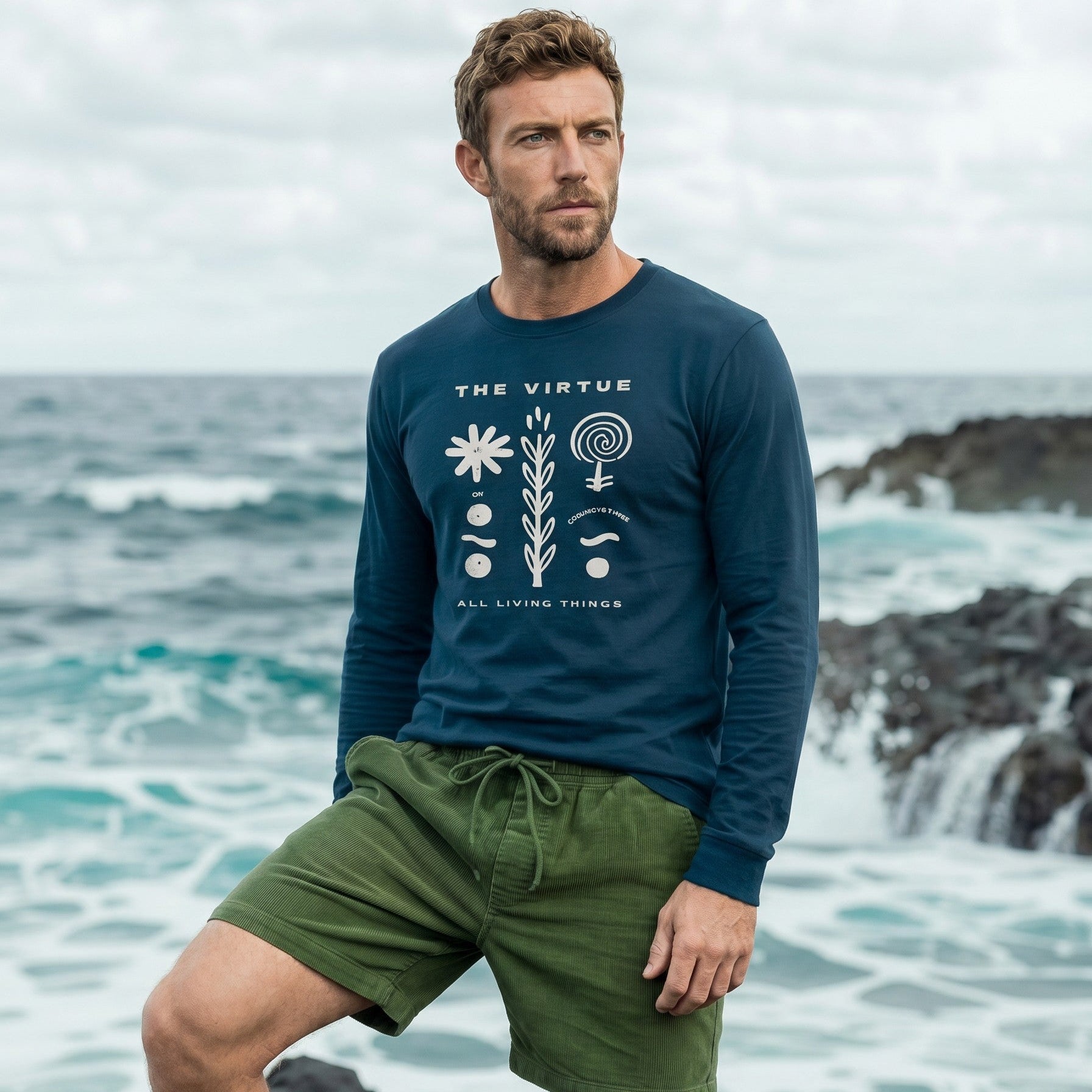 Man wearing a blue shirt and green shorts standing on a rocky beach with ocean waves in the background.